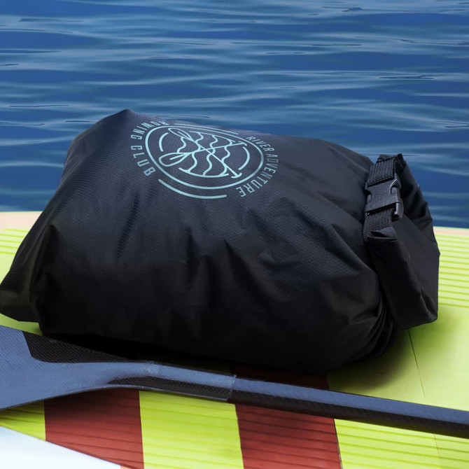A black lightweight dry bag with a logo, resting on a paddleboard beside calm water.
