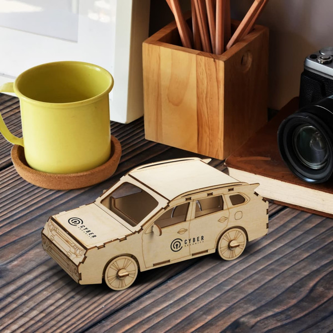A wooden SUV model in natural wood finish with a logo, placed on a wooden surface near a cup and camera.
