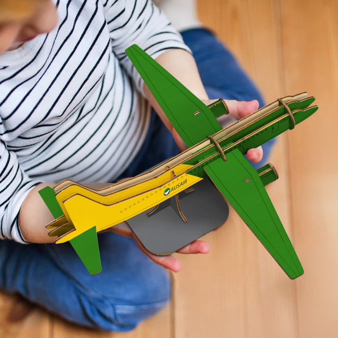 A wooden model of a jet plane in green and yellow, held by a child. The model is mounted on a grey base.