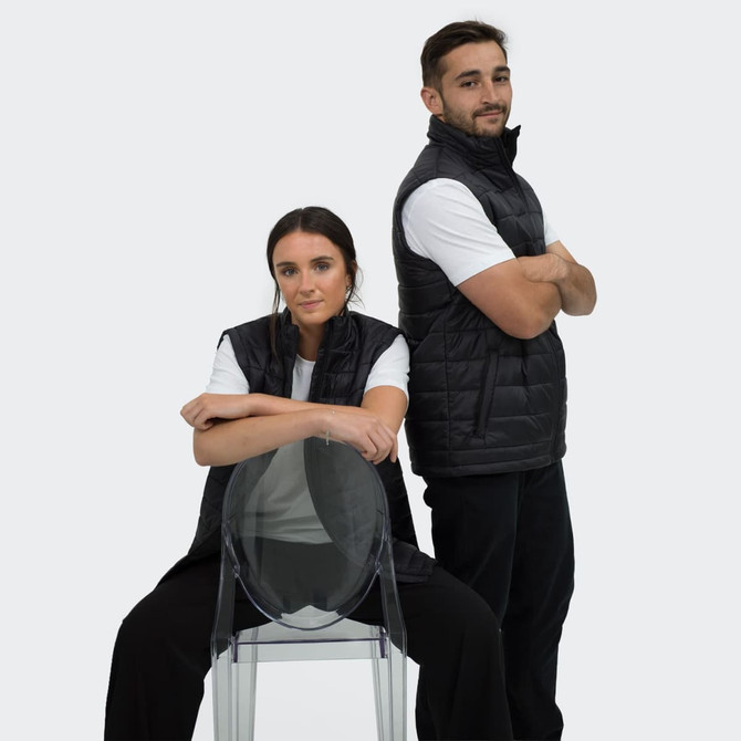 Black puffer vest worn by two models, one seated and one standing, both in white t-shirts and casual pants.