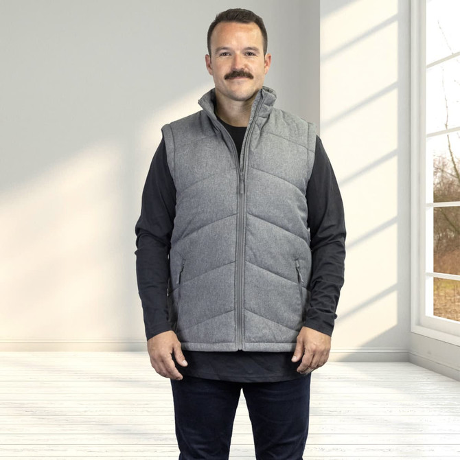 A man stands in a well-lit room wearing a grey puffer vest over a black long-sleeve top. The vest has a logo.