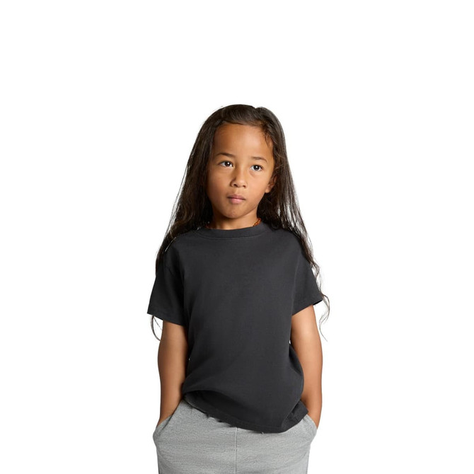 A black kids' heavy faded tee with short sleeves, worn by a child with long hair, standing with hands in pockets.