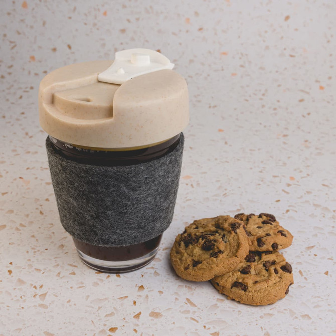 A reusable coffee cup in grey with a lid, featuring a felt band, accompanied by chocolate chip cookies.