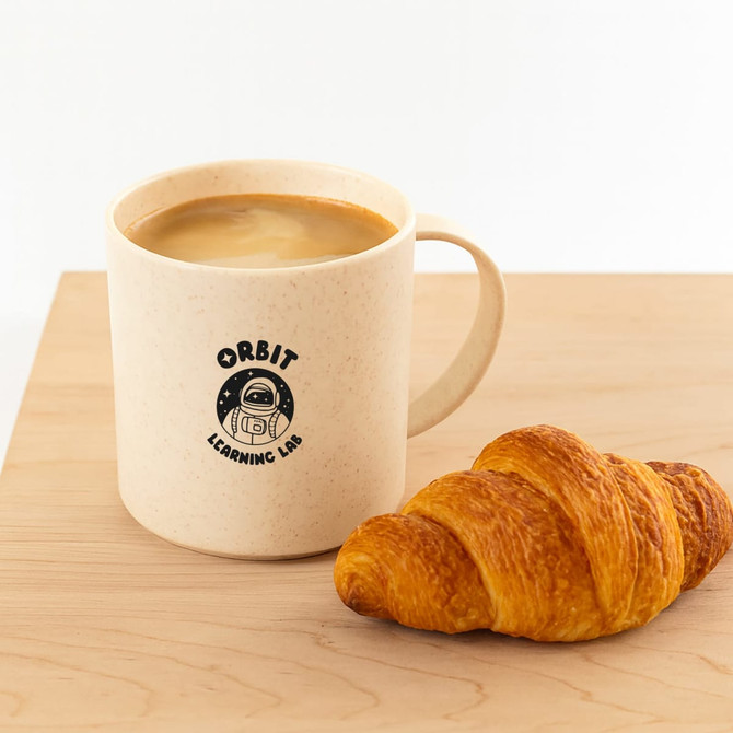 A reusable beige mug with a handle, featuring a logo, alongside a flaky croissant on a wooden surface.