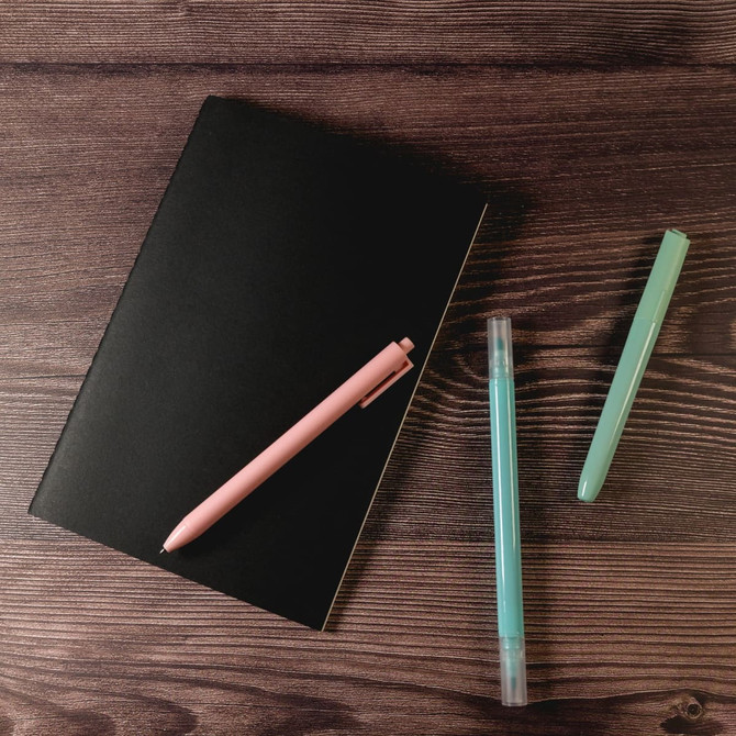 A black notebook lies on a wooden surface, accompanied by a pink pen and two green highlighters.