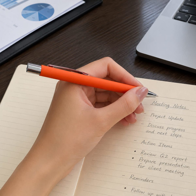 An orange metal pen held above a notepad with handwritten meeting notes, placed on a wooden desk near a laptop.