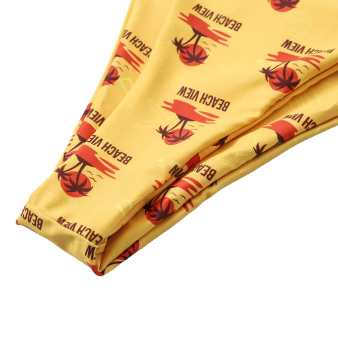 A yellow bikini bottom featuring a beach view print with palm trees and sun motifs. It includes a logo.