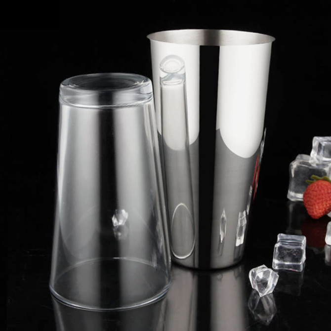 A cocktail shaker set featuring a transparent plastic cup and a shiny stainless steel shaker, placed on a dark surface with ice and fruit.