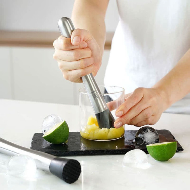 A stainless steel cocktail muddler in use, with lime halves and ice in a glass on a black slate surface.