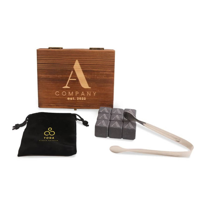 A wooden box with a logo, containing grey whiskey stones, a pair of tongs, and a black velvet pouch.