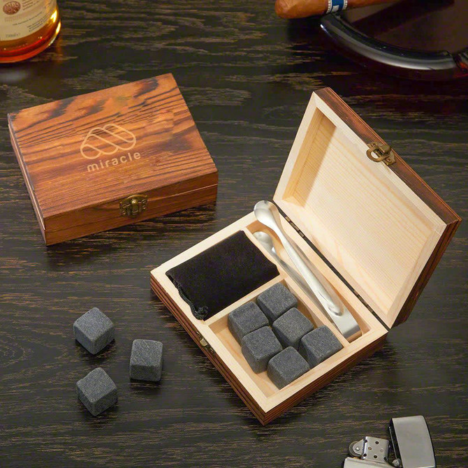 A wooden box containing grey whiskey stones, a pouch, and two metal tongs, featuring a logo on the lid.