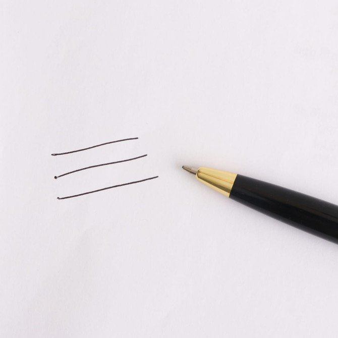 A black plastic click pen with a gold tip beside three lines drawn on white paper. The pen features branding.