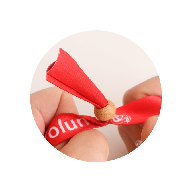 A red RPET fabric wristband with a bamboo toggle, held between two hands. The wristband features a logo.