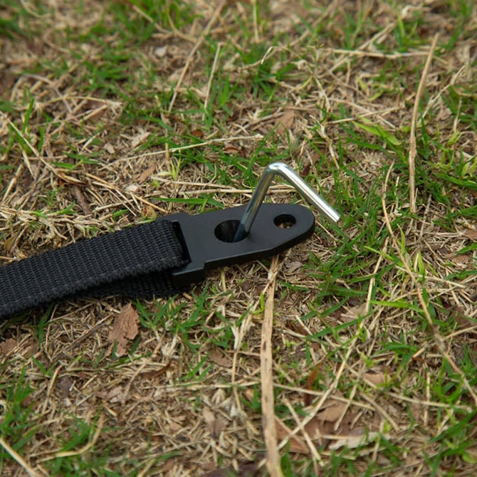 A black strap with a metal anchor pin on grass, designed for securing outdoor equipment.