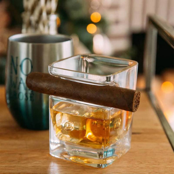 A whiskey glass with a square design, filled with amber liquid, holds a cigar on the rim. The background features blurred lights.