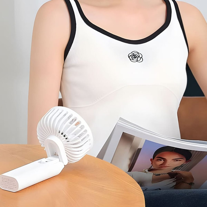 A white handheld power bank folding fan sits on a table beside a person in a white top, with a magazine nearby.