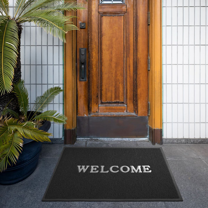 A black floor mat with a logo in front of a wooden door, surrounded by greenery.