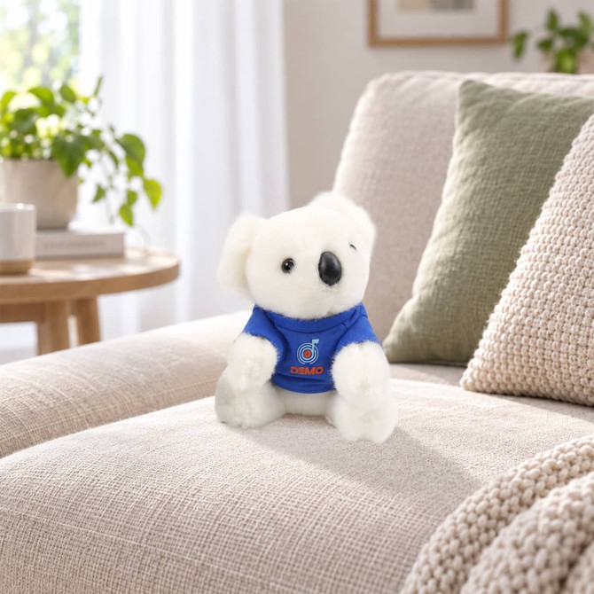 A soft white koala plush toy wearing a blue outfit, sitting on a beige sofa in a cozy living space.