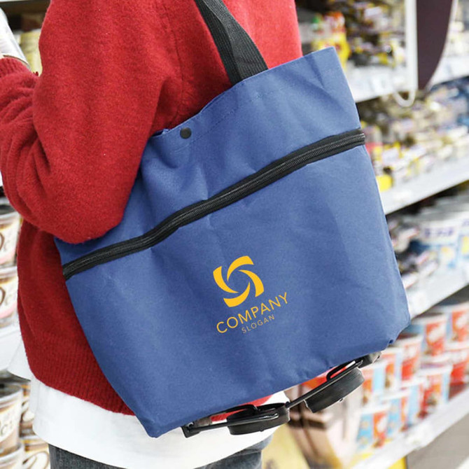 A blue collapsible shopping trolley bag with a black strap and a logo, held by a person in a red sweater.