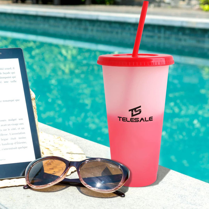 A pink and white colour changing cup with a red lid and straw, placed beside sunglasses and an e-reader by a pool.