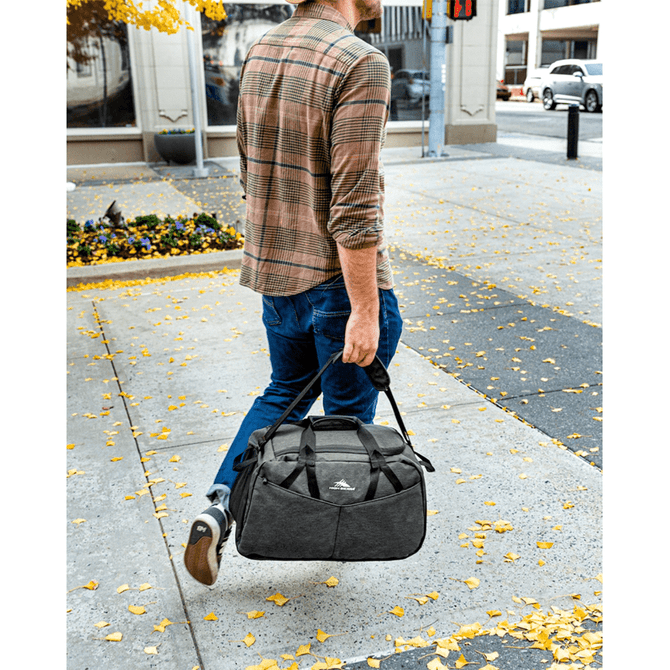 A man walks outdoors carrying a large grey duffel bag with black accents and a logo.