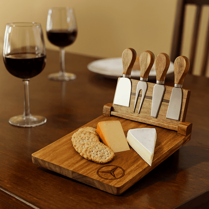 A wooden cheeseboard set featuring four stainless steel knives, cheese, and crackers, with two glasses of red wine in the background.