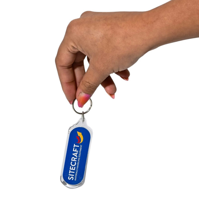 A hand holds an oval acrylic keychain with a silver metal ring and a blue logo on one side.