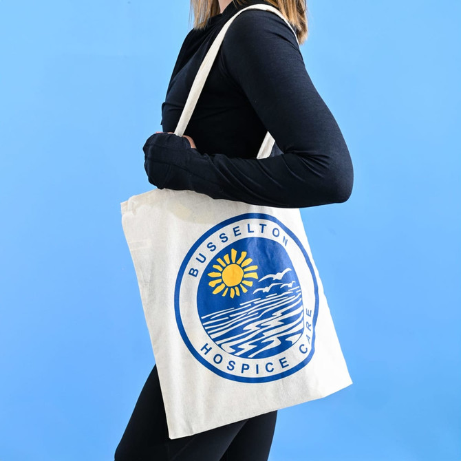 A cream Calico tote bag with a circular design featuring a sun and waves, held by a person against a blue background.