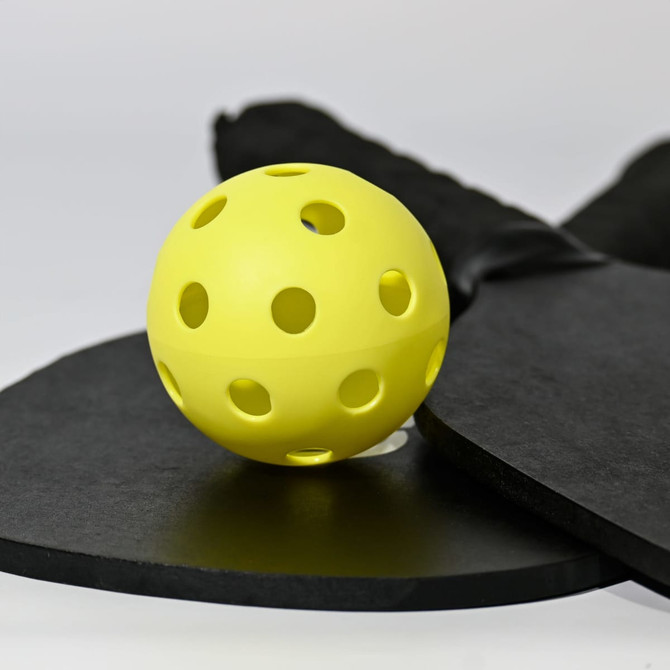 A yellow pickleball with holes, resting on a black paddle and a round black surface, featuring a logo.