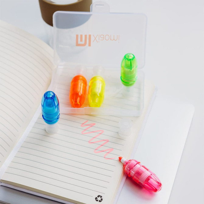 A transparent case containing four highlighters in blue, orange, yellow, and pink, placed on a notepad with lines.