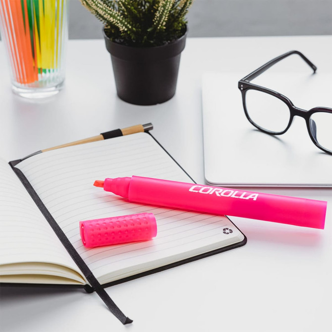 A pink jumbo highlighter with a cap rests on an open notebook beside a plant and glasses.
