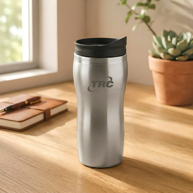 A sleek silver travel mug with a black lid and a logo, placed on a wooden table next to a plant and notebook.