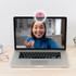 A ring selfie light on a laptop screen, illuminating a smiling woman in a blue top. The laptop is on a wooden desk.
