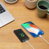 A black 10000mAh power bank with a logo beside a smartphone, on a wooden desk with a coffee cup.