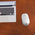 A white wireless mouse sits on a wooden table beside a laptop with a black keyboard. The mouse has a logo.