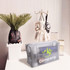 A grey nylon mesh cosmetic bag sits on a table next to a potted plant and decorative items, featuring a logo.