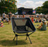 A folding chair in black with a mesh back sits on grass, facing a crowd at an outdoor event with a stage.
