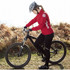 A woman in a red lightweight jacket and helmet is riding a black mountain bike in a grassy area.