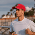 A man in a white shirt wears a bright red cap with a logo, set against a coastal background.