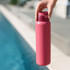 A drink bottle in a vibrant pink colour, being held by a hand near a swimming pool. It features a logo.