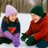 Two children in winter attire wearing knitted gloves, one in purple and the other in green, playing in snow.