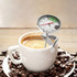 A silver instant read dial thermometer is placed in a steaming coffee cup, surrounded by coffee beans.
