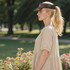 A woman in a beige dress wearing a patterned brown visor stands in a garden with flowers. The visor features a logo.