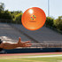An orange frisbee is being thrown, featuring a logo in the centre. The background shows bleachers in a sports field.