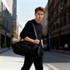 A black convertible bucket backpack duffel bag is held by a man standing on a city street. The bag features a logo.