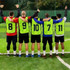 Five players wearing brightly coloured training bibs in green, red, and blue, facing away with arms raised on a field.