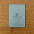 A soft PU leather notebook in a muted green colour with a logo on the cover, placed on a wooden surface.