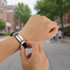 A metal strap bracelet with a black band is being adjusted on a wrist, set against a blurred urban background.