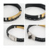 A black leather bracelet featuring a gold bar with branding. The design includes white stitching and a metallic clasp.
