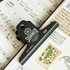 A metal binder clip in black holds music sheets and decorative paper on a surface, featuring a logo.